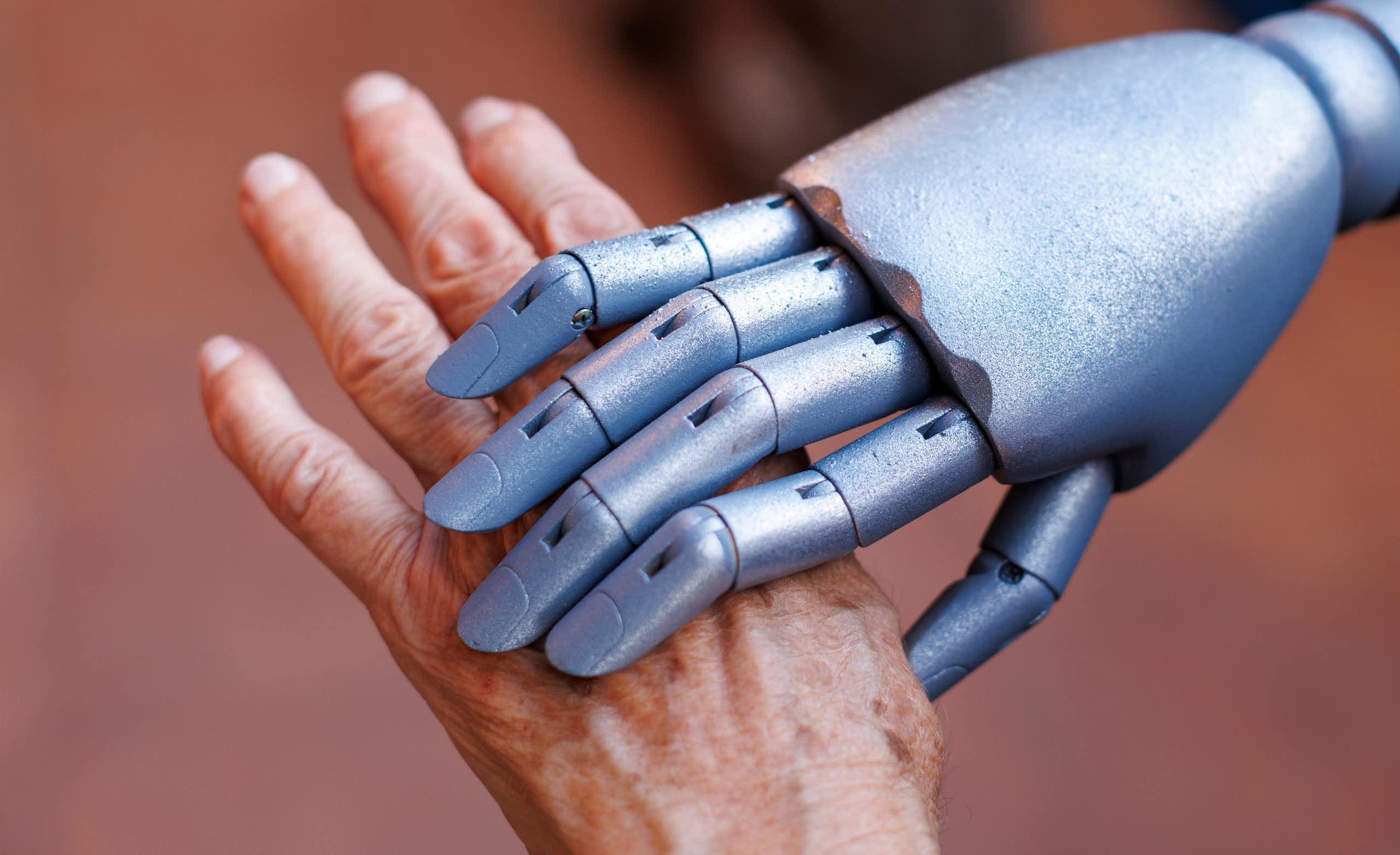 Uma mão robótica prateada segura a mão de uma pessoa idosa.