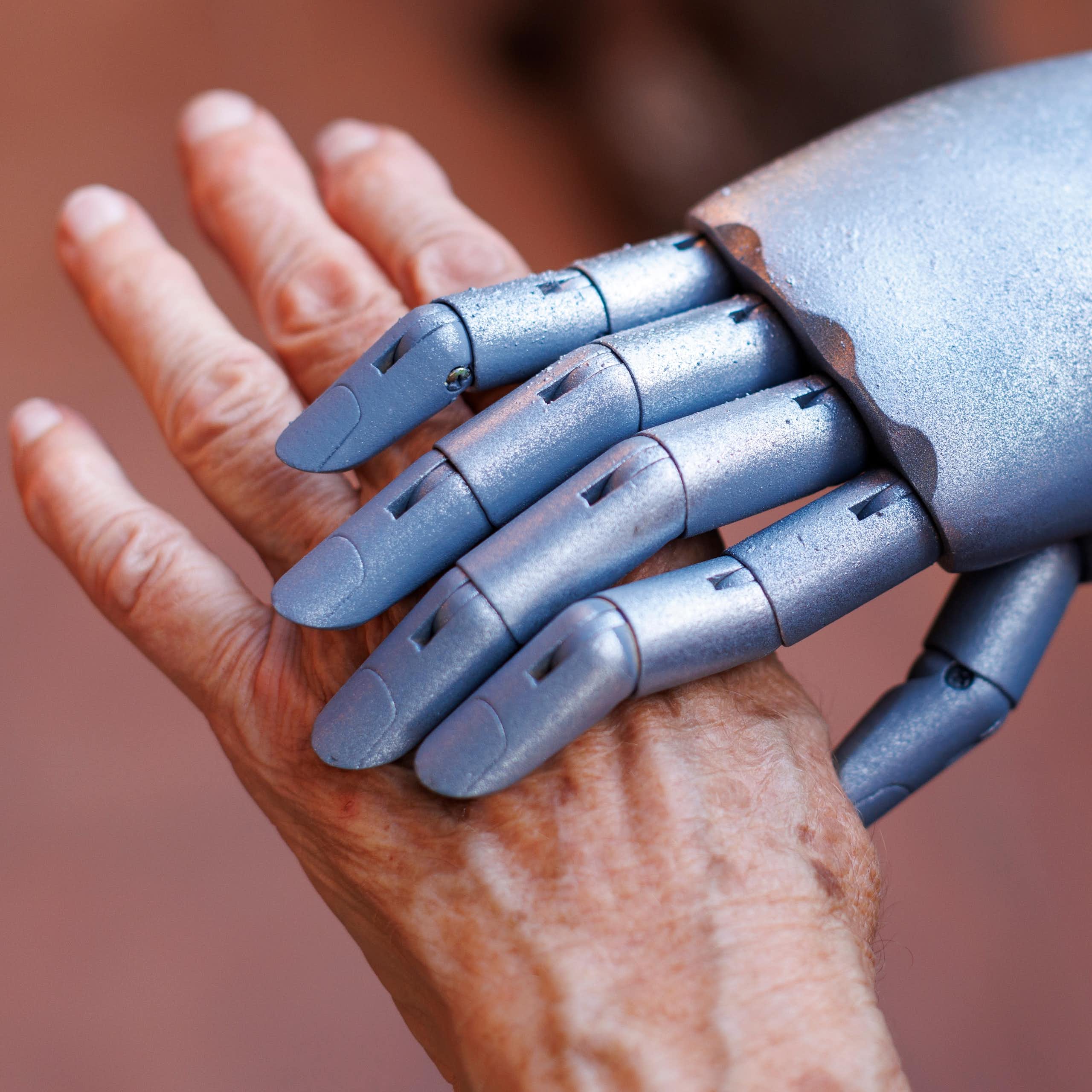 Uma mão robótica prateada segura a mão de uma pessoa idosa.