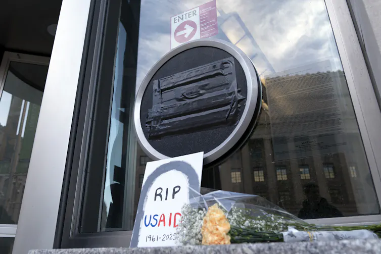 A round plaque blacked out with tape, with a hand-lettered sign reading RIP USAID and a bunch of flowers