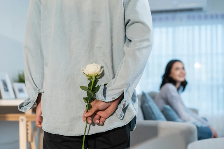 A man from the neck down with a white rose behind his back as a woman sits looking at him in the background.