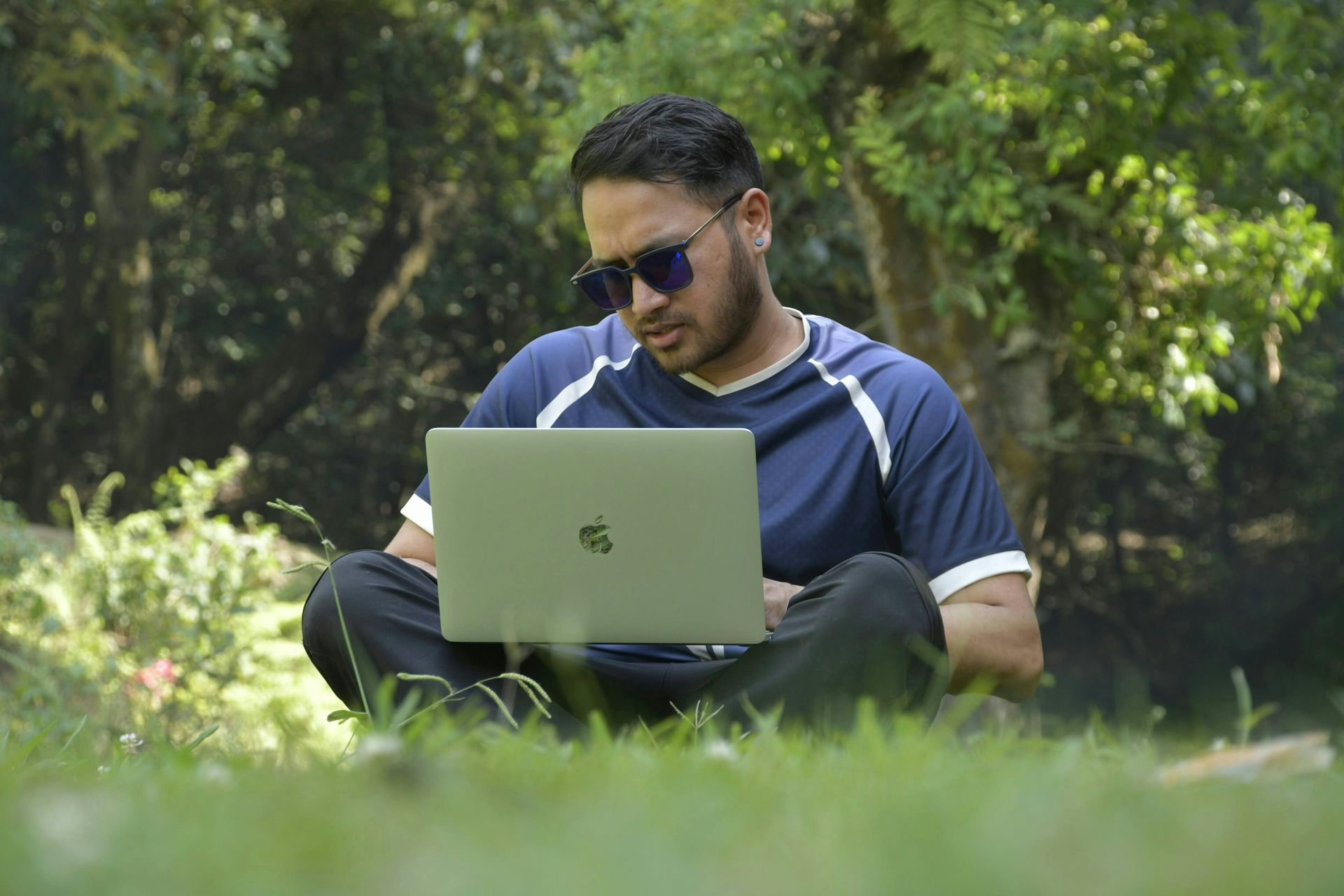 A man in sunglasses sits in a park working on a laptop.