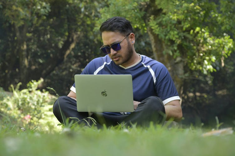 A man in sunglasses sits in a park working on a laptop.