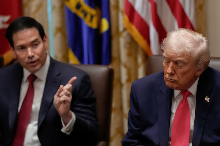 An older man with fluffy white-ish blond hair and orange-hued skin appears to be asleep as a younger man next to him talks.
