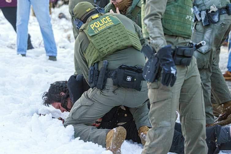 A man in a green army uniform with a vest that says 'Border Patrol' stands over a person who lies face down in the snow.