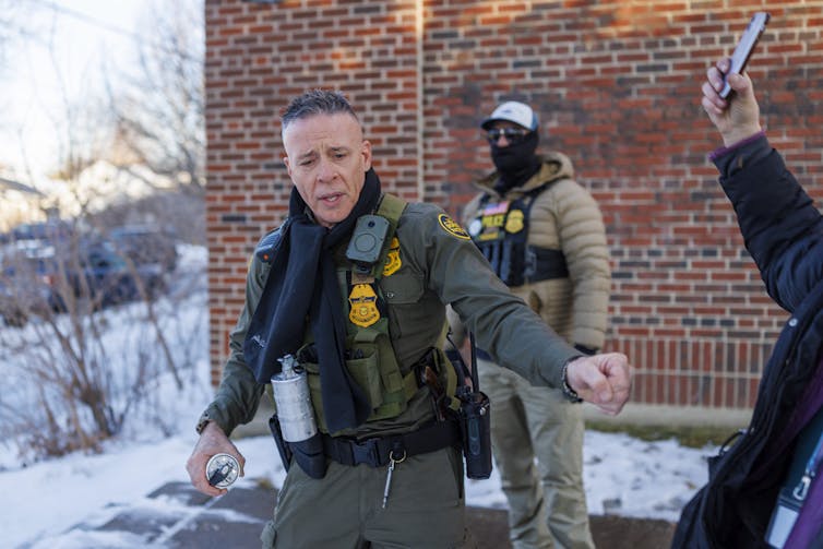 Federal immigration enforcement close to colleges disrupts attendance, traumatizes scholars and damages their instructional efficiency 1 A man wearing a green vest and pants stands near another man also in a green uniform, in front of a red brick building.
