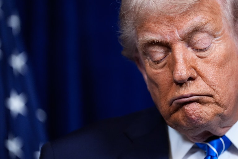 An elderly man with fluffy white hair and orange-hued skin appears to nod off.