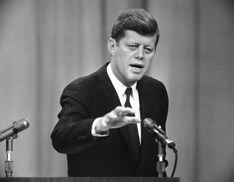 A black-and-white photo shows a youthful man with thick light brown hair talking at a lectern behind a microphone.