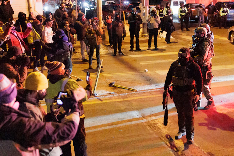 on a city street at night a crowd of people use their phones to record several men wearing tactical vests and gas masks