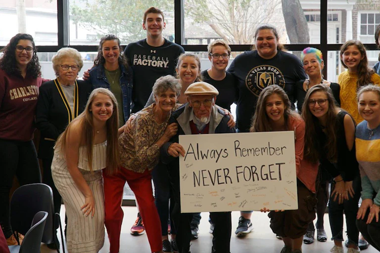 An elderly man holding a poster that says 'Always remember, never forget' poses with two rows of young people.