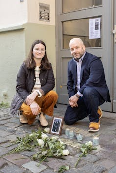 Bearing witness after the witnesses are long gone: Learn how to deliver Holocaust schooling house for a brand new technology 2 A young woman and a man in a blue suit kneel by a small memorial installed on a sidewalk of paving stones.