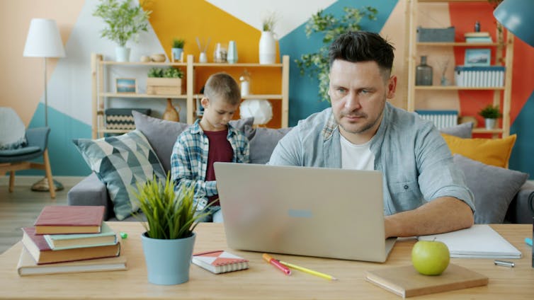 A father works on a laptop, his son sits on a couch in the background