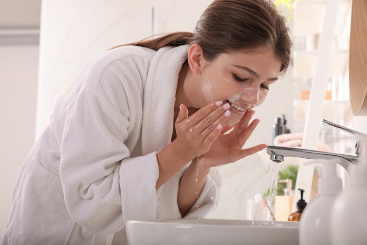 A woman wearing a white bath robe uses a cleanser to wash her face in a bathroom sink.