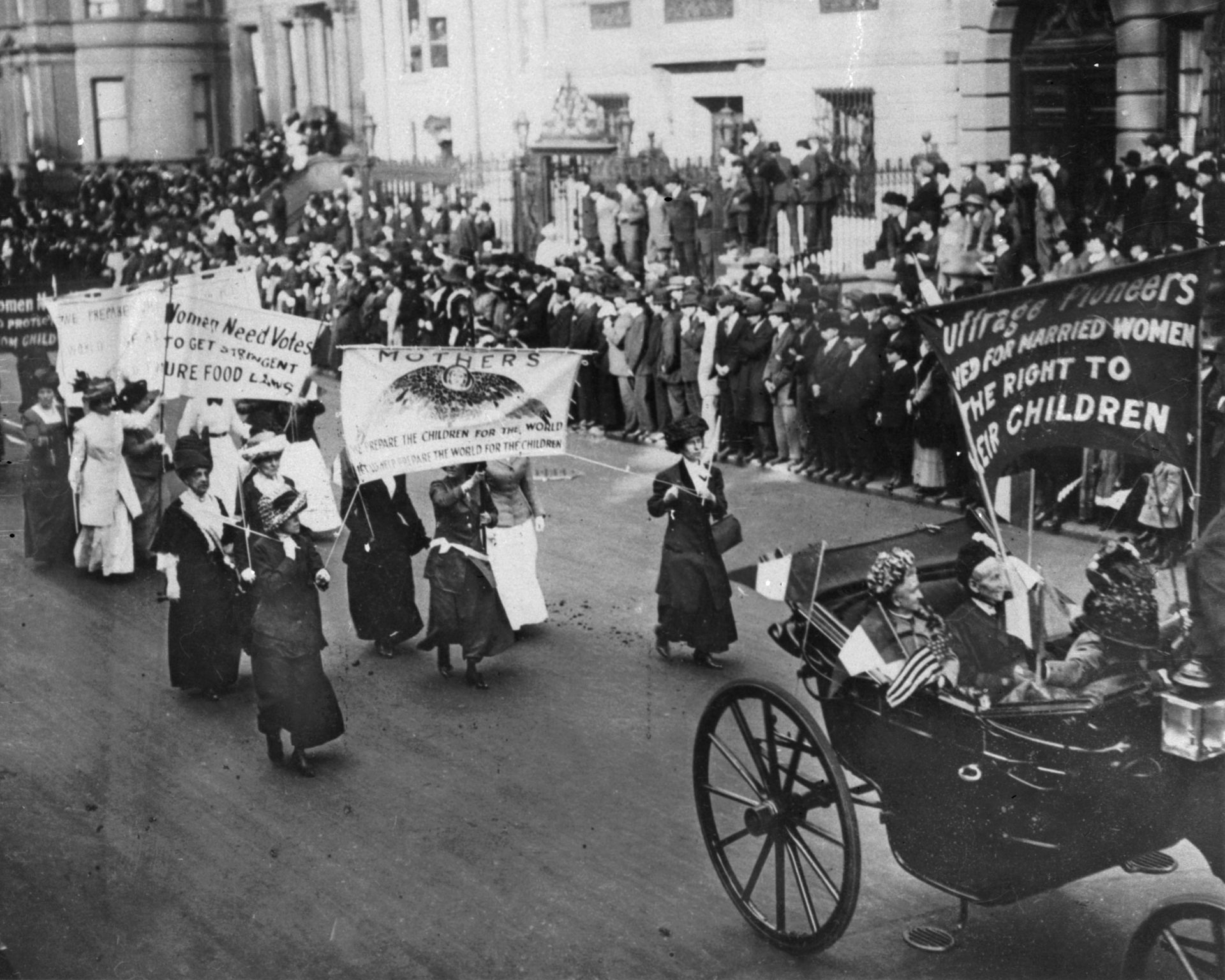 a black-and-white photo shows women in long coats and hats marching and waving banners