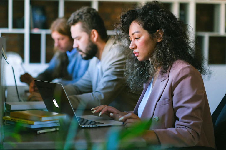 Woman sits at a computer with two colleagues next to her. She is wearing a white t-shit and pink suit jacket.