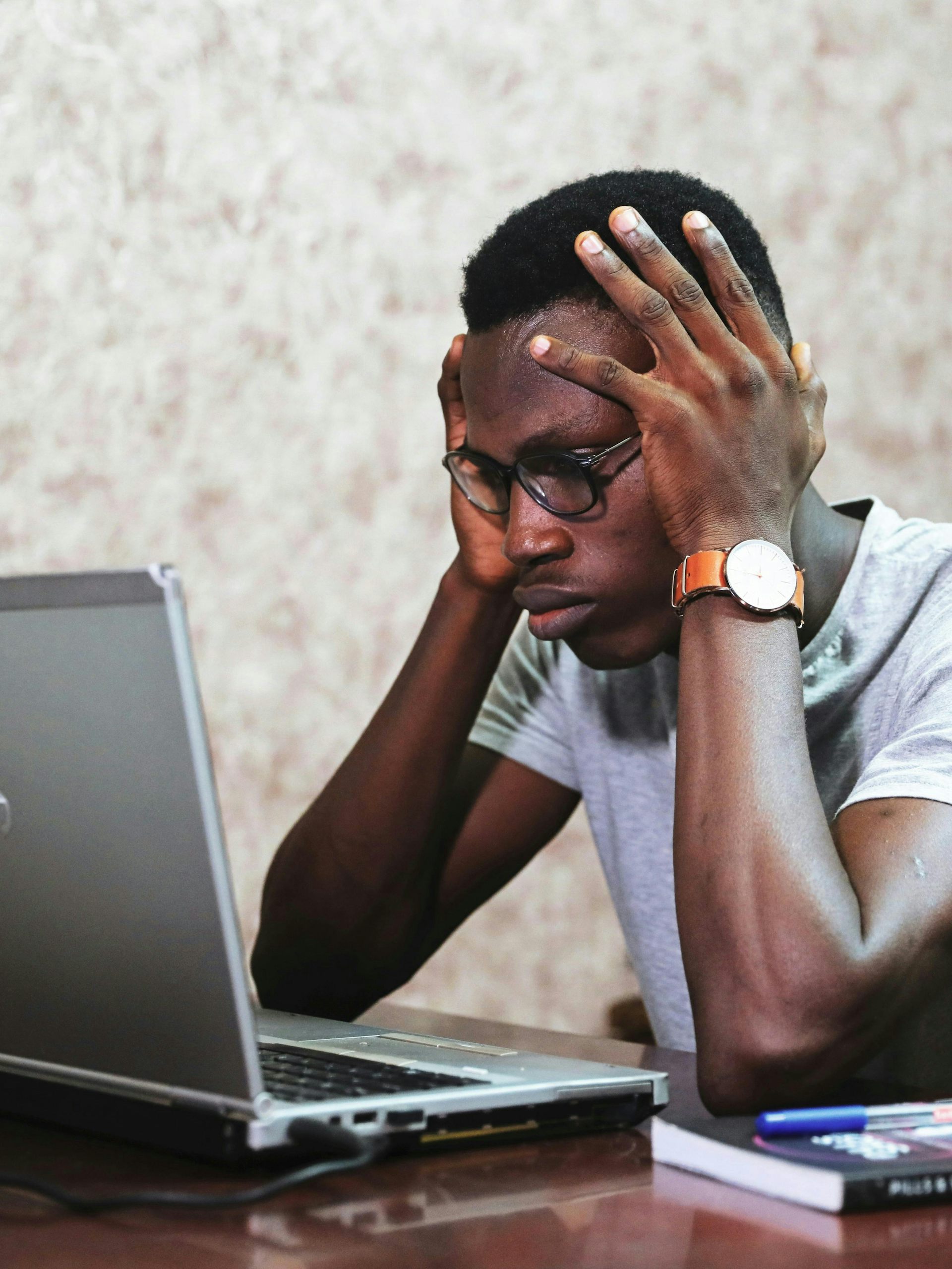 A man sits at a computer looking stressed, holding his head in his hands.