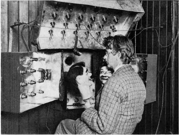 John Logie Baird sits with two handheld puppets behind a system for transmitting moving image, March 1925.