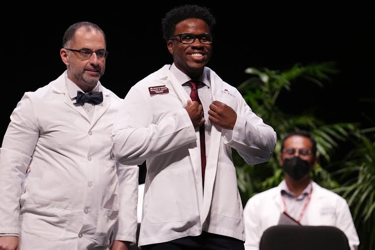 younger man smiles putting on a short white doctor's coat with two older men behind