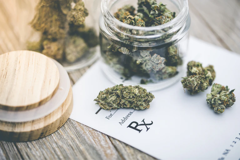 Clear, open bottle of marijuana buds surrounded by loose buds on top of a paper with Rx label.