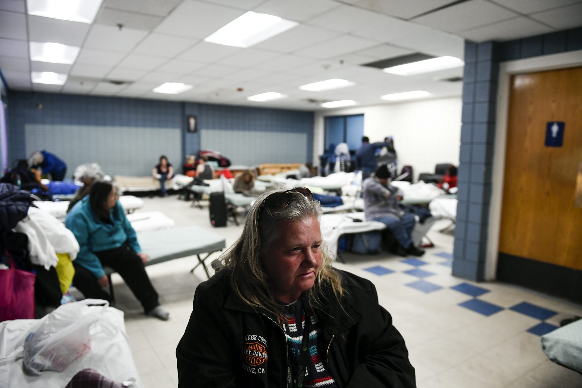 A woman sits in a room of other people on cots.
