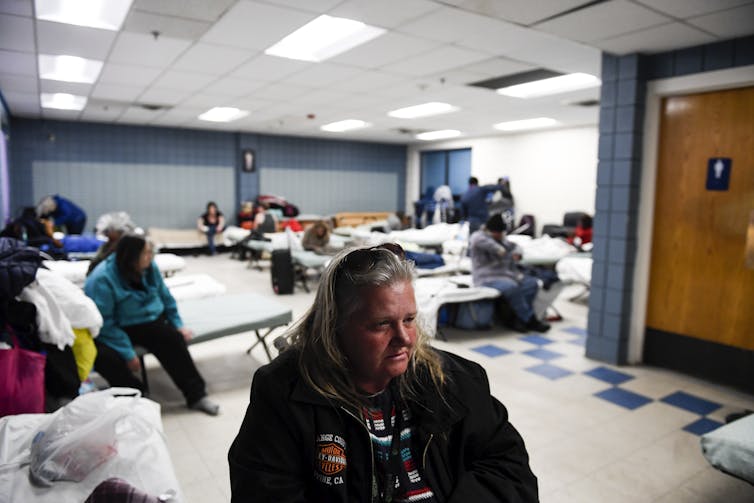 A woman sits in a room of other people on cots.