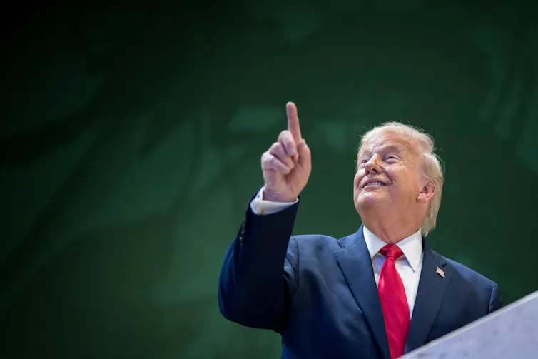 A grinning older man with fluffy white-blond hair smiles as he looks up and points.