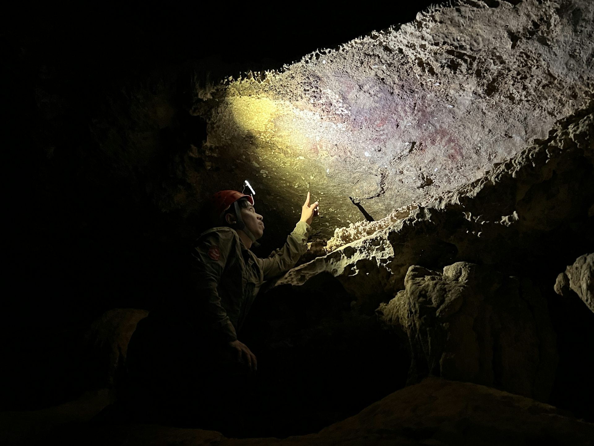 Um homem em uma caverna escura apontando para antigas pinturas rupestres na superfície rochosa.