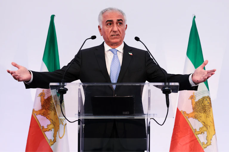 A gray-haired man in a suit standing at a lectern in front of some flags, with his arms spread.