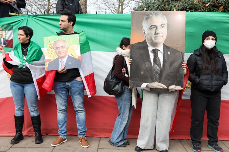 Protesters holding enlarged photos of Reza Pahlavi as they stand on a street, some of them wearing flags around their shoulders.