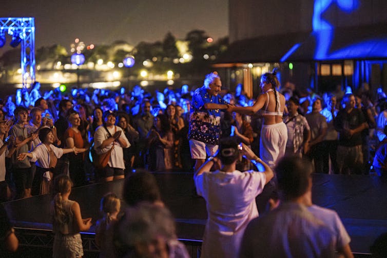 A large group of people dance together outside.