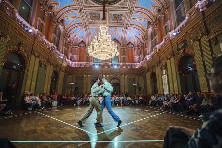 Two men dance in an ornate hall.