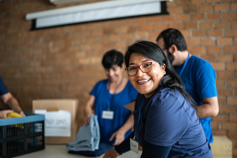 A Latina woman smiles as she and other people in blue shirts pack boxes with donated goods.