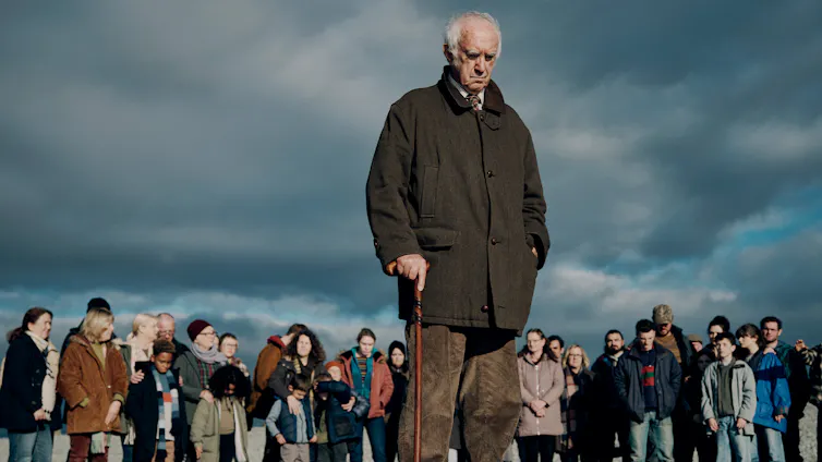 Pryce leans on a walking stick, crowd of people in the background