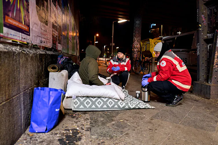 Two people in red coats crouch on a sidewalk, speaking with someone in a green jacket seated atop blankets.