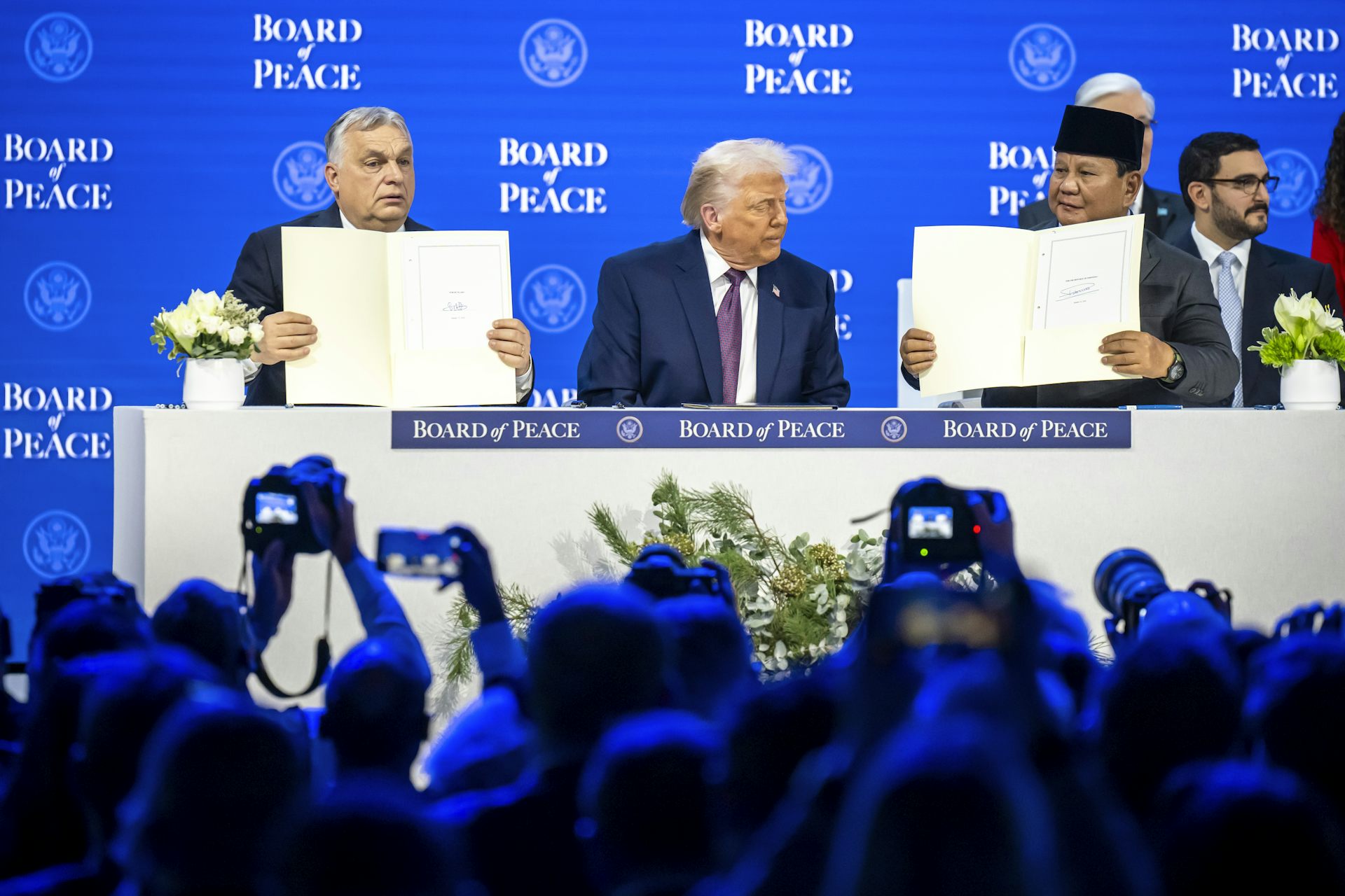 Viktor Orbán, Donald Trump and Prabowo Subianto sitting at a table in front of a crowd, holding up documents they've signed.