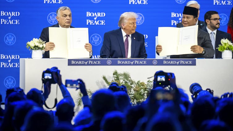 Viktor Orbán, Donald Trump and Prabowo Subianto sitting at a table in front of a crowd, holding up documents they've signed.