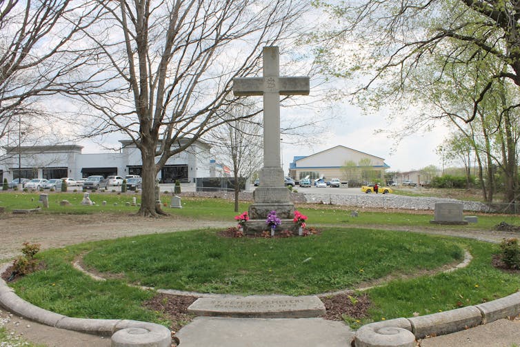 A large stone cross stands over a grave in a circular, grass-covered memorial, with a few trees in the background.