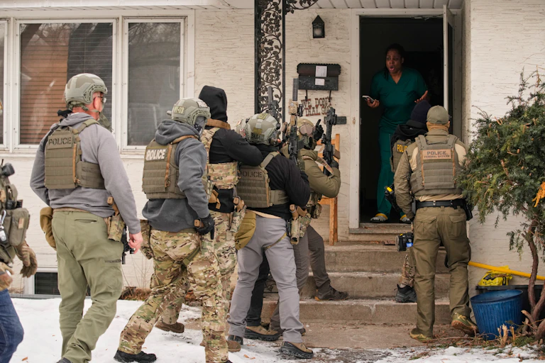 A woman standing in a house doorway, looking out at a group of people dressed in police regalia.