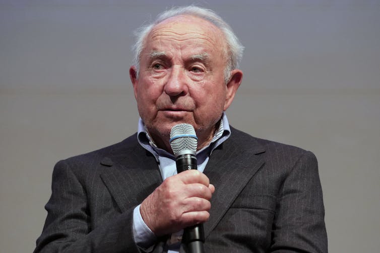 An old man with a round face and silver hair holds a microphone while speaking at an event.
