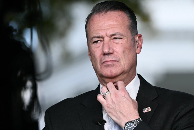 A dark-haired man looking grim and fiddling with his white-collared shirt.