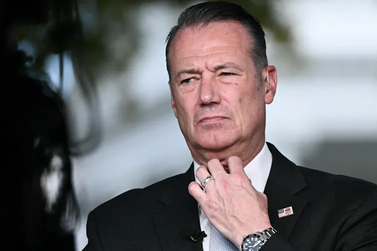A dark-haired man looking grim and fiddling with his white-collared shirt.
