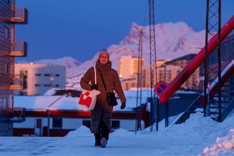 A woman clutches her bag as she walks along a snowy street, with snow-covered mountains in the background.