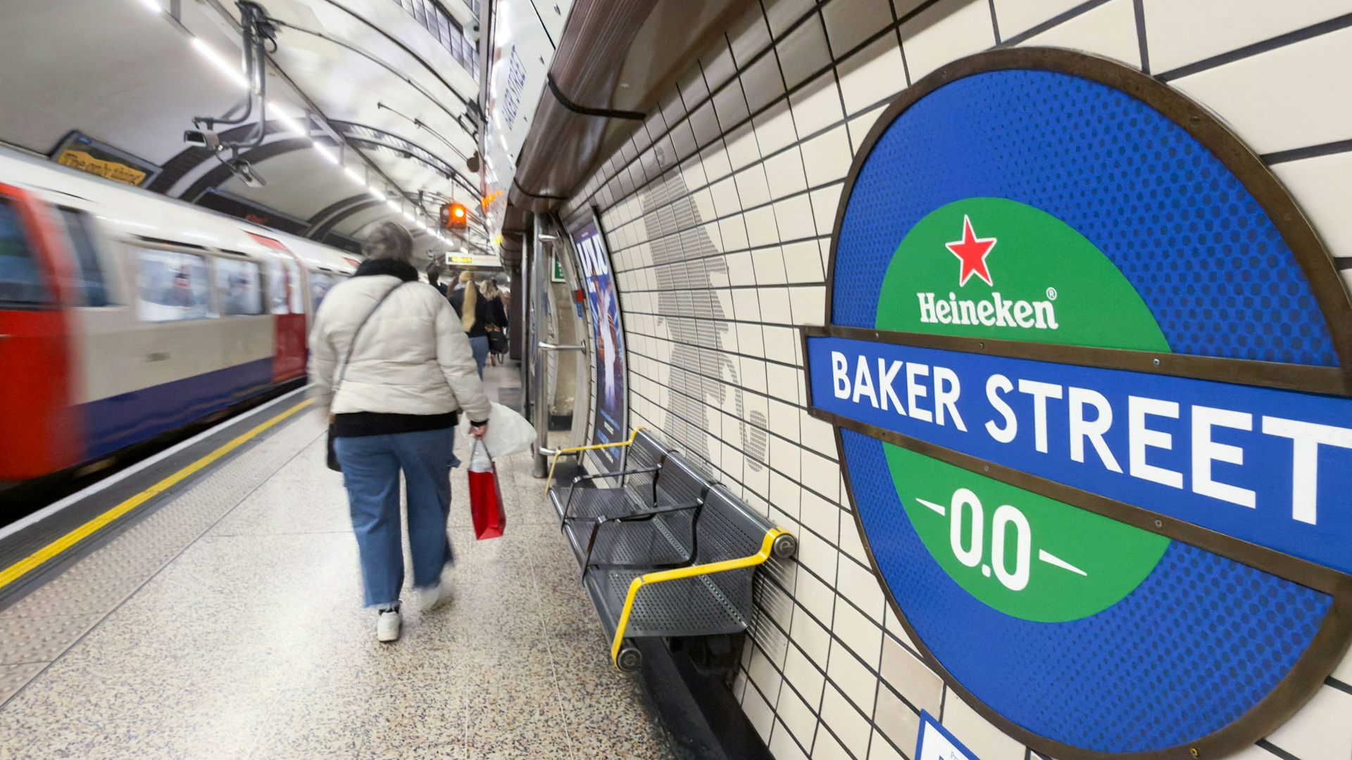 A passenger walks past a Heineken-branded roundel displaying "Baker Street -00-" as a train waits at the platform, Baker Street Underground station.