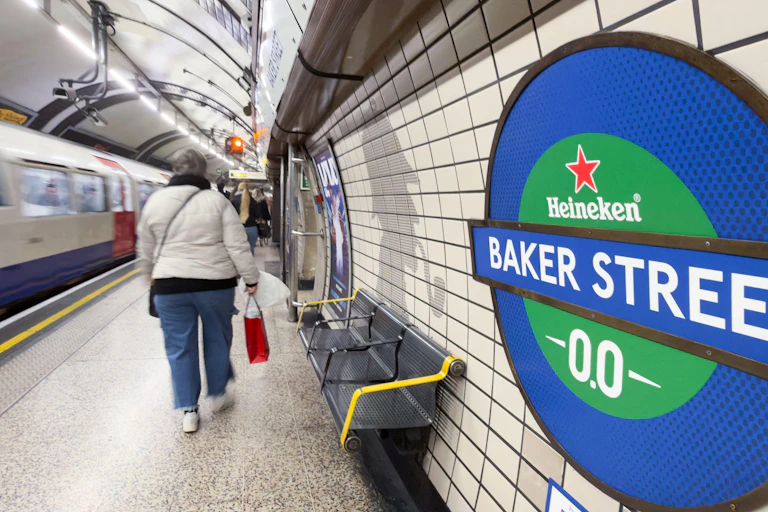 A passenger walks past a Heineken-branded roundel displaying "Baker Street -00-" as a train waits at the platform, Baker Street Underground station.