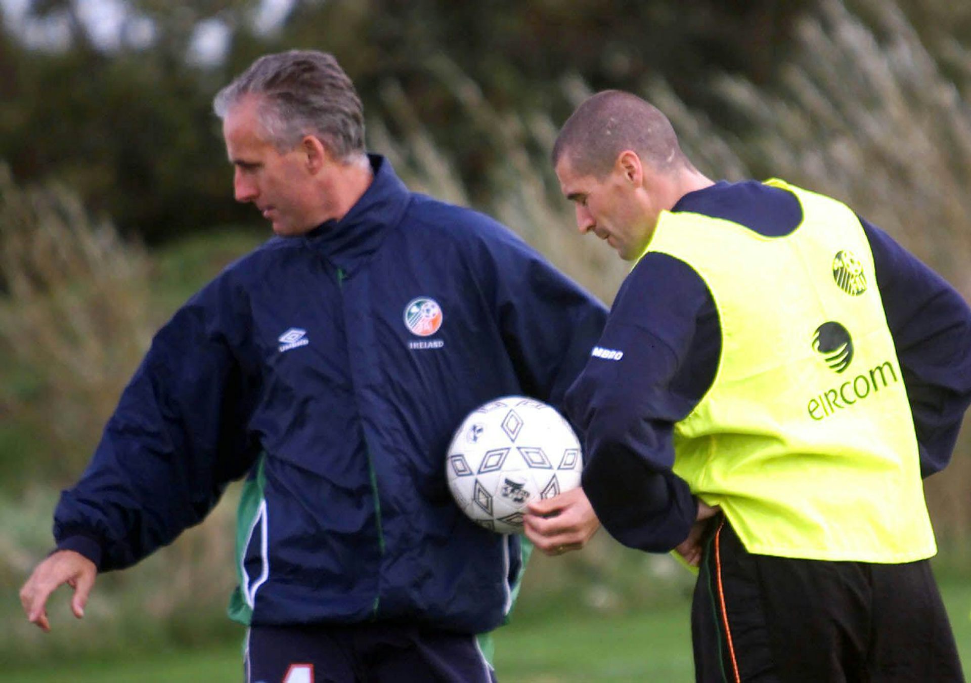 An older grey-haired man with a football in his hand and a young player in a training vest.