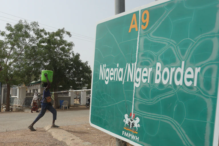 Man carrying a container on his head walks near a sign board saying Nigeria/Niger Border