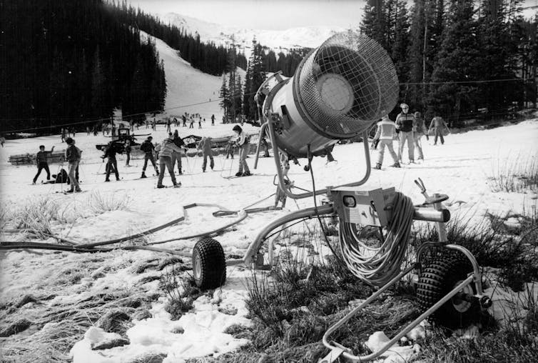 Las estaciones de esquí de Colorado recibieron algunas nevadas bienvenidas debido a la tormenta invernal Fern, pero no las suficientes para revertir un invierno seco y cálido. 2 Una fotografía en blanco y negro muestra una máquina de nieve en primer plano y esquiadores al fondo en una montaña.