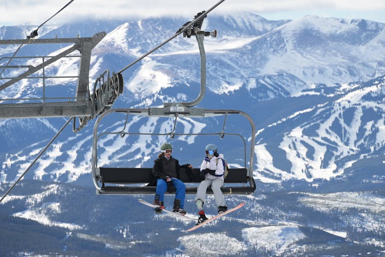 Two people in winter clothes sit on a ski lift with a ski resort with sparse snow visible in the background.