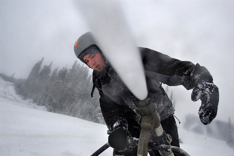 A man in snow gear stands behind a machine blasting a stream of snow into the air.