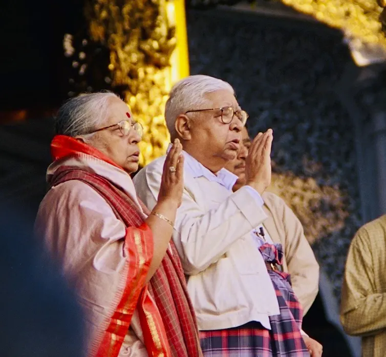 A couple -- the woman wearing a sari with a red border and the man in a white shirt -- raise their hands in a gesture of blessing while standing.