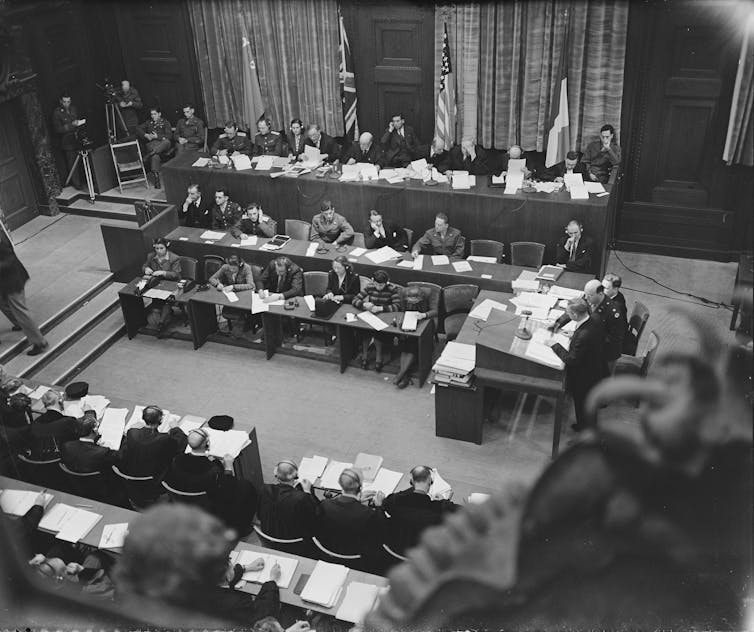 A courtroom scene shows several people seated in three rows, with national flags displayed behind them and additional rows of seated attendees visible in front.